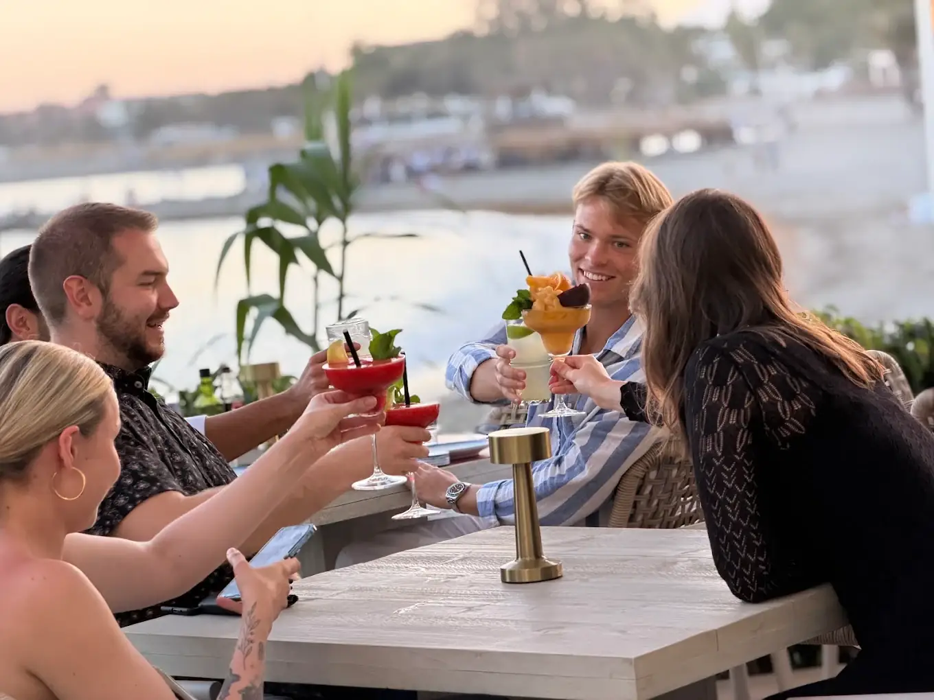 Friends raising cocktails at a terrace table at sunset, the beach behind them