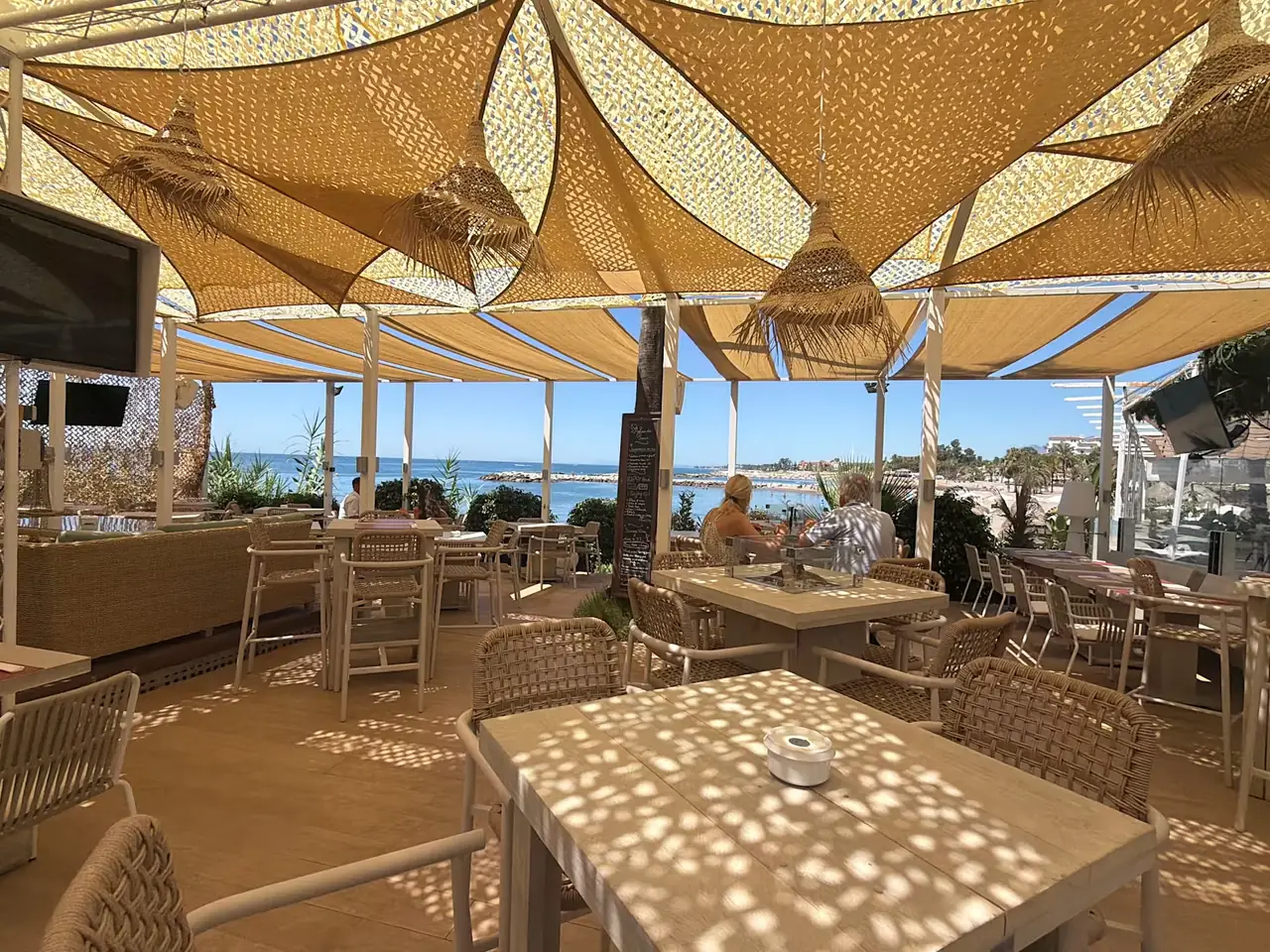 Dappled light through the shade sails across the terrace tables, the beach in the distance