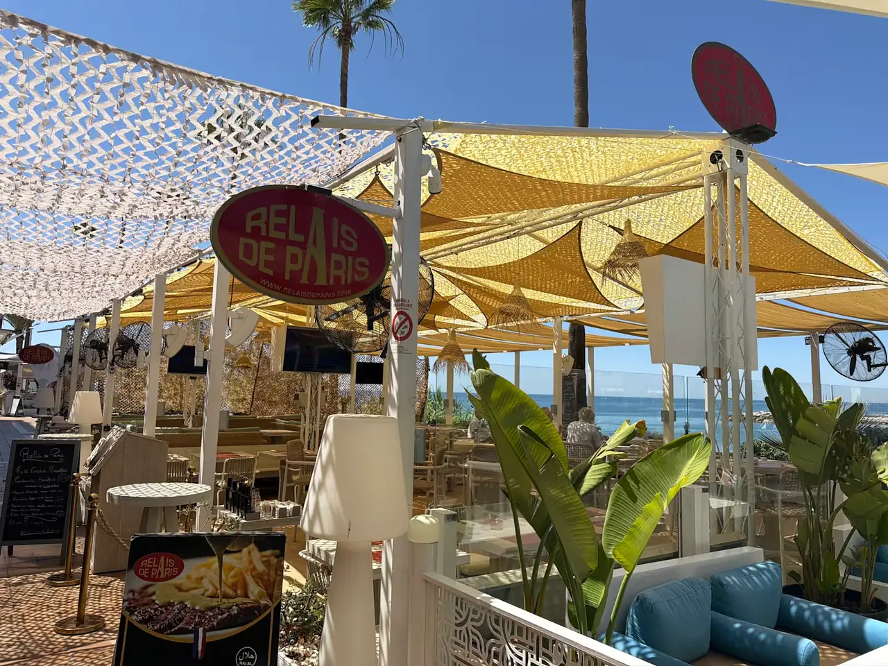 The red oval Relais de Paris sign above the terrace, palm trees and blue sky behind
